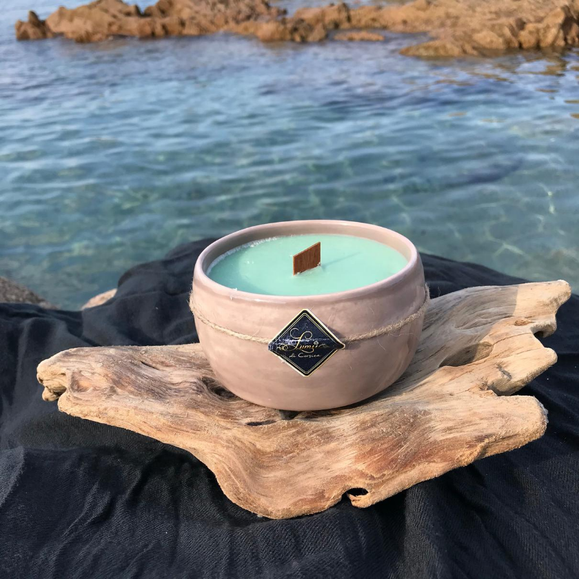 Céramique artisanale Corse, avec un bougie à l'interieur de couleur verte, une mèche en bois marron et un étiquette bleu ou il est noté Lumii di Corsica. Elle est posé sur un bois à flotter et une nappe noir. Les rocher et la mer sont en arriere plan.