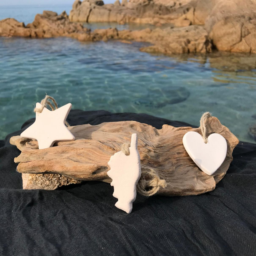 Un lot de pierre à parfumer en céramique, composé d'une étoile, d'une Corse et d'un coeur. Chacun à une ficelle et ils sont posés sur un morceau de bois à flotter avec une nappe noir. En arrierre plan la mer te la montagne Corse.